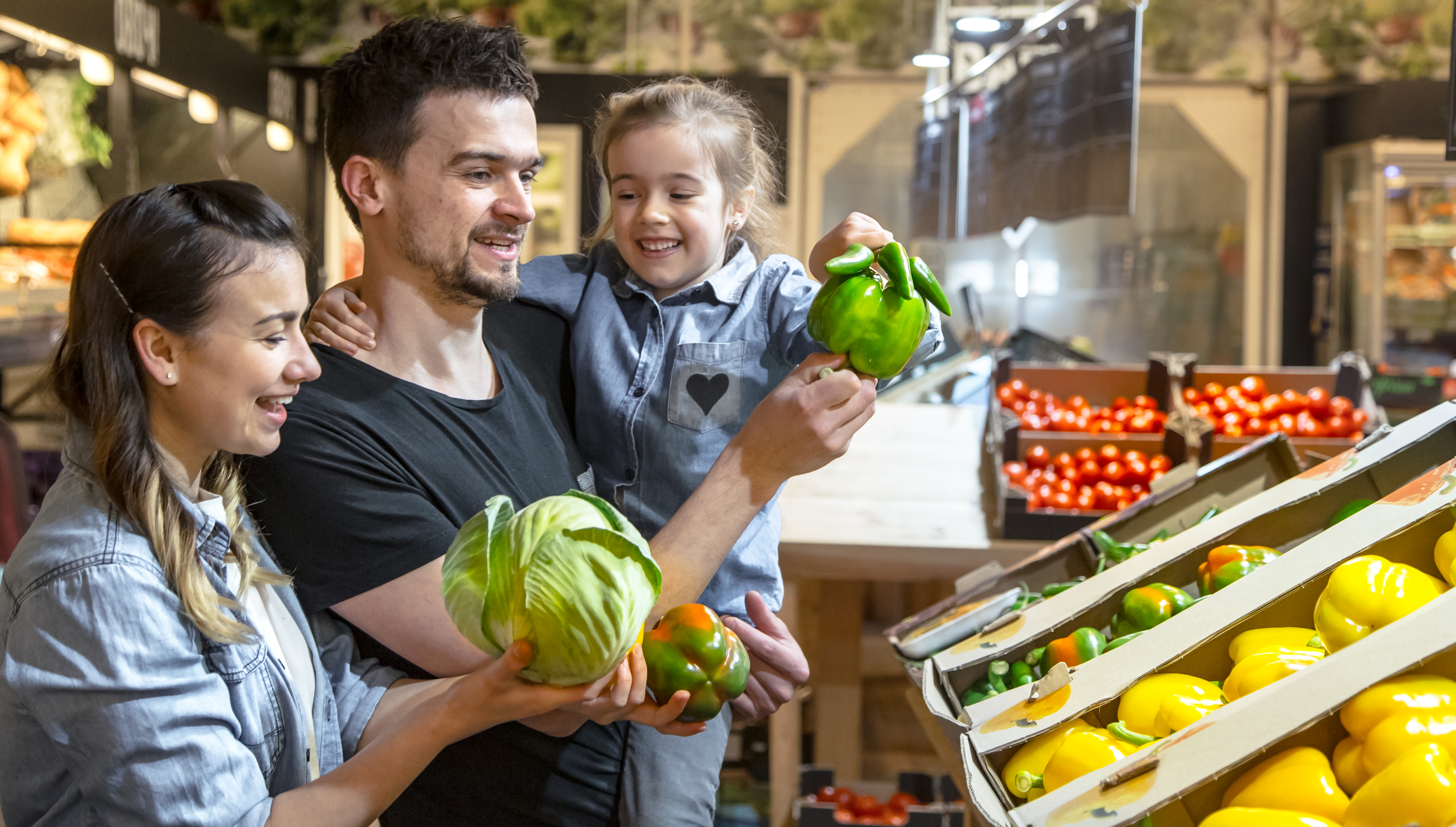 happy husband and wife with kid buys vegetables cheerful family of three choosing bell pepper and greens in vegetable department of supermarket or market